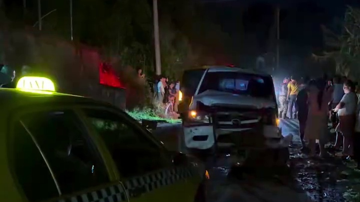 Pick-up blanco con el frente destruido junto a un paredón, de noche, con vecinos alrededor y un taxi en primer plano