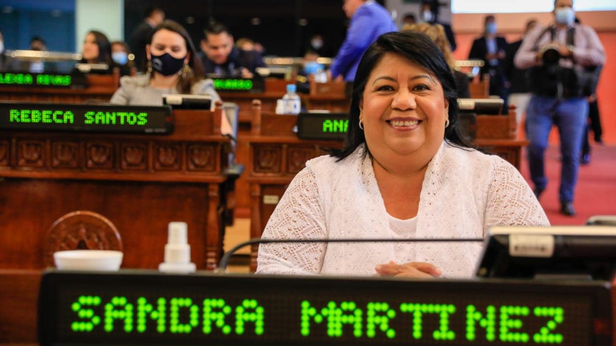 Sandra Yanira Martínez sonríe sentada en su curul de la Asamblea Legislativa, con blusa blanca